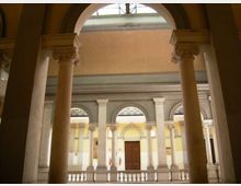 Cortile interno di un edificio storico con porticato ad archi e colonne in pietra con capitelli decorati. Sullo sfondo si vedono una facciata con finestre ad arco, una porta centrale in legno e un lucernario in alto.