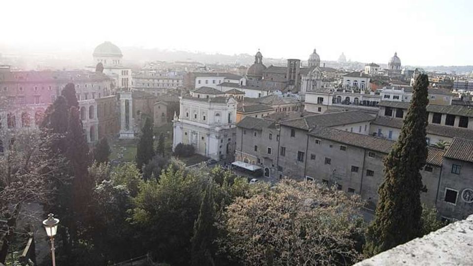 Vista panoramica su un centro storico con edifici in muratura e tetti in tegole, interrotta da cupole e campanili sullo sfondo. In primo piano si vedono alberi alti (cipressi) e una terrazza in pietra che affaccia su un’area verde.