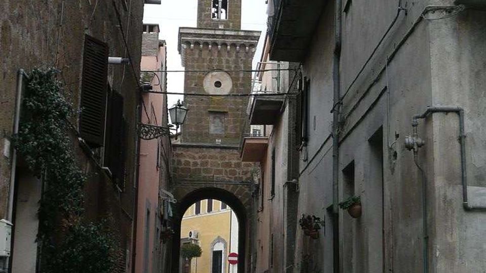 Vicolo stretto in un centro storico, fiancheggiato da alti edifici in muratura con balconi, tubature esterne e vasi di piante lungo il muro. In fondo si vede un arco in pietra che conduce a una piazzetta e una torre campanaria con merlature e finestre ad arco.