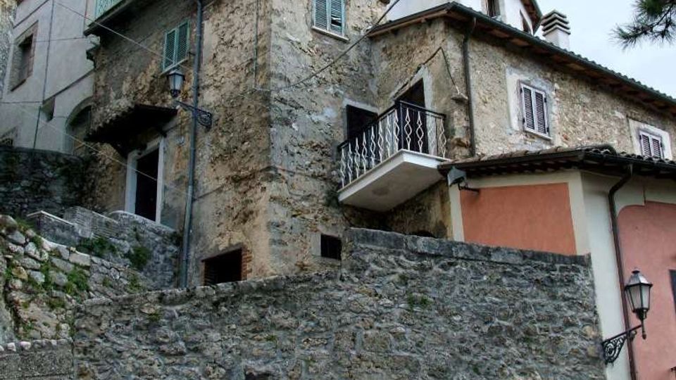 Edifici residenziali su pendio con facciate in pietra e intonaco, tetti in tegole e piccoli balconi con ringhiere metalliche. In primo piano si vede un alto muro di contenimento in pietra con arco e portone, affiancato da una lanterna a muro.