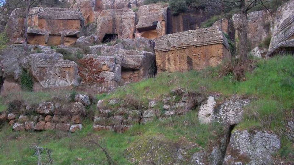 Necropoli rupestre su pendio erboso, con tombe e camere funerarie scavate nella roccia e grandi blocchi di pietra squadrati. Sullo sfondo si vedono pareti rocciose con aperture rettangolari e alberi sparsi.