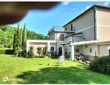 Casa di campagna a due piani con facciata chiara, balcone con ringhiera metallica e portico coperto arredato. Davanti c’è un ampio prato verde con piante ornamentali e lampioni da giardino, sullo sfondo colline boscose e cielo azzurro.