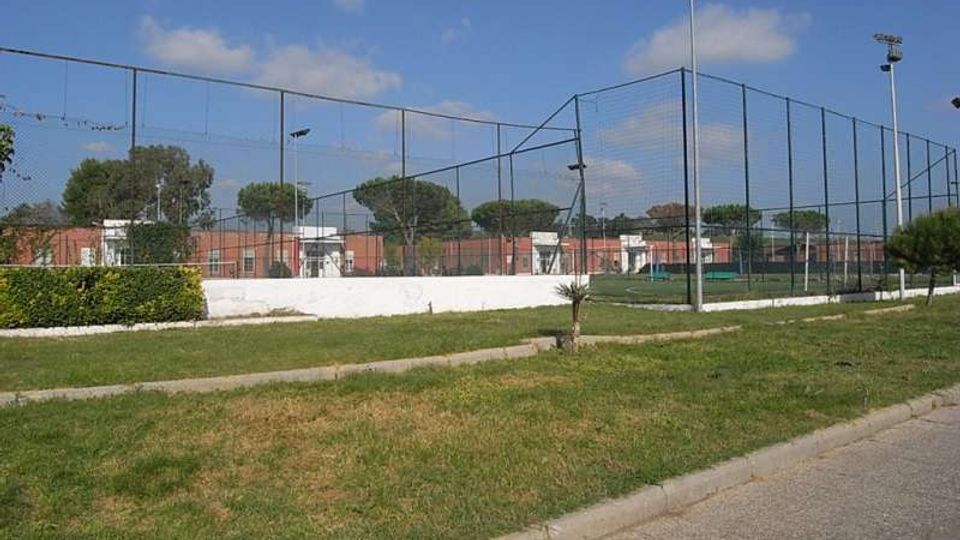 Campo sportivo all’aperto recintato con alte reti metalliche e pali di illuminazione, circondato da un prato e un vialetto asfaltato. Sullo sfondo si vedono alberi e edifici bassi con cielo parzialmente nuvoloso.