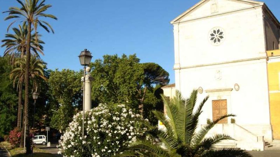 Facciata di una piccola chiesa intonacata di bianco con rosone circolare e croce sul frontone, affacciata su una piazza. In primo piano ci sono palme, un cespuglio fiorito e un lampione, con alberi sullo sfondo sotto un cielo sereno.