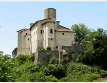 Castello in muratura chiara su una collina, con torre cilindrica centrale e tetti in coppi rossi. In primo piano si vede un muraglione di pietra con arco e vegetazione fitta tutt’intorno.