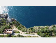 Veduta aerea di una costa rocciosa con vegetazione mediterranea e mare blu intenso. In basso si vede una strada costiera e un piccolo edificio rettangolare vicino al bordo della scogliera.