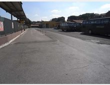Ampio piazzale asfaltato di un deposito autobus, con pensiline e recinzioni sul lato sinistro e diversi autobus parcheggiati sulla destra. Sullo sfondo si vedono edifici bassi e alberi sotto un cielo parzialmente nuvoloso.