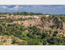Panorama di un borgo con case in pietra su un altopiano, affacciato su una scarpata rocciosa a strapiombo. In primo piano si vede una valle verde con alberi e vegetazione fitta sotto un cielo parzialmente nuvoloso.