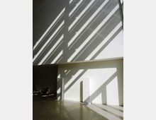 Atrio interno moderno con pareti bianche e pavimento in grandi lastre, attraversato da lunghe ombre diagonali create da una vetrata o lucernario. Sul lato sinistro si intravedono sedute e un’area più in ombra.