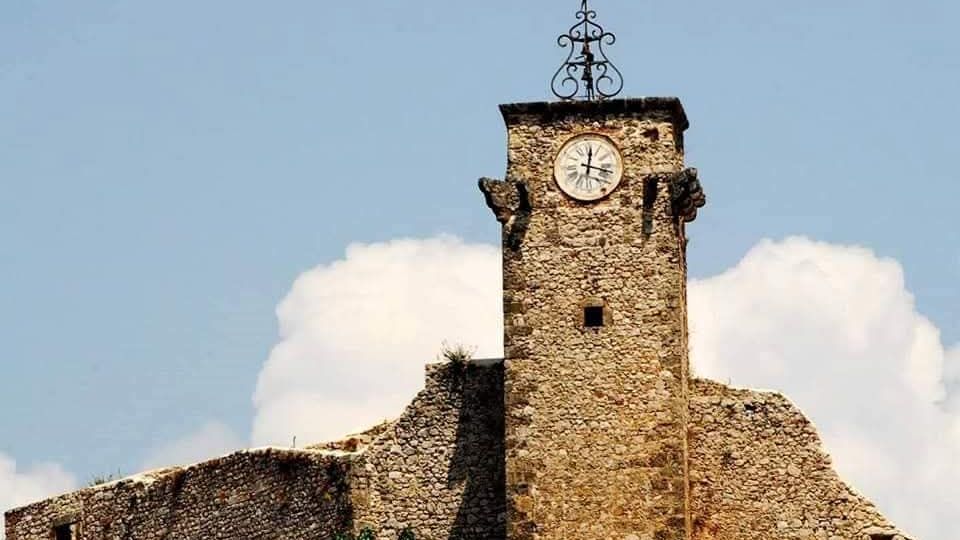 Torre campanaria in pietra con orologio circolare sulla facciata e croce in ferro battuto in cima, integrata in un complesso di mura e tetti in pietra. Sullo sfondo si vede il cielo azzurro con nuvole e vegetazione alla base della struttura.