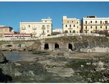 Veduta di un lungomare con edifici chiari a più piani affacciati sulla costa, separati da una staccionata bianca. In primo piano si vedono scogli e piattaforme rocciose con piccole grotte alla base di un muretto in pietra.
