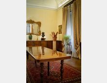 Sala da pranzo in stile classico con lungo tavolo in legno su tappeto orientale rosso, boiserie scura e grande specchio dorato sopra una credenza. Ampia finestra con tende pesanti e velo bianco, con pianta su piedistallo, soffitto alto decorato e arredi d’epoca.