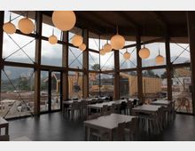 Interno di una sala ristorante moderna con grandi vetrate a tutta altezza incorniciate da travi in legno e tiranti metallici, con lampade sferiche sospese dal soffitto. Tavoli e sedie disposti in file su pavimento scuro, con vista verso un’area esterna e montagne sullo sfondo.