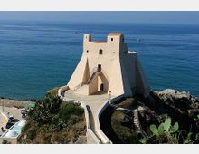 Fortezza costiera in pietra chiara con mura inclinate e piccola torre, arroccata su uno sperone roccioso affacciato sul mare. Una passerella e una scalinata conducono all’ingresso, con il litorale e l’orizzonte marino sullo sfondo.