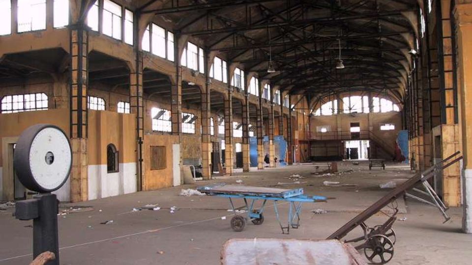 Interno di un grande capannone industriale dismesso, con soffitto alto a capriate metalliche e lunghe file di finestre che illuminano una navata centrale. Pavimento in cemento con detriti sparsi e alcuni carrelli e attrezzature abbandonate in primo piano.