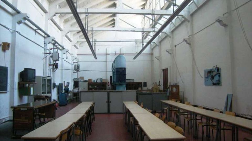 Ampio laboratorio/aula tecnica con lunghi tavoli e sedie disposti in file, pavimento rosso e attrezzature industriali lungo le pareti. Soffitto alto con capriate bianche e lucernari longitudinali che illuminano l’ambiente.