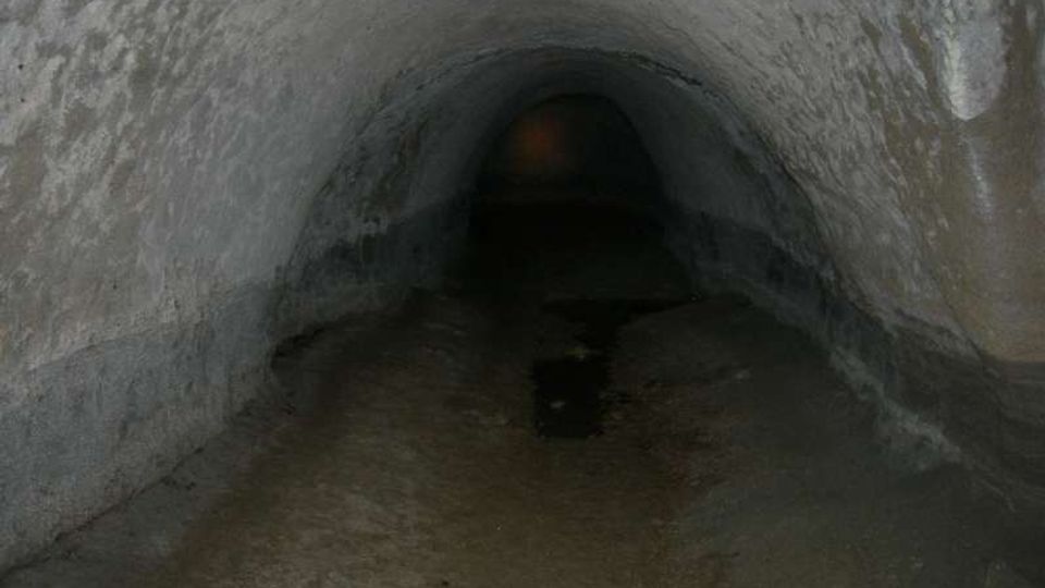 Interno di un tunnel sotterraneo con volta a botte in pietra grezza e pavimento umido. Il corridoio si restringe e scompare nell’oscurità verso il fondo.