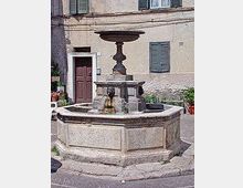 Fontana in pietra a più vasche con colonna centrale, situata in una piccola piazza lastricata davanti alla facciata di un edificio con porta in legno e persiane verdi.