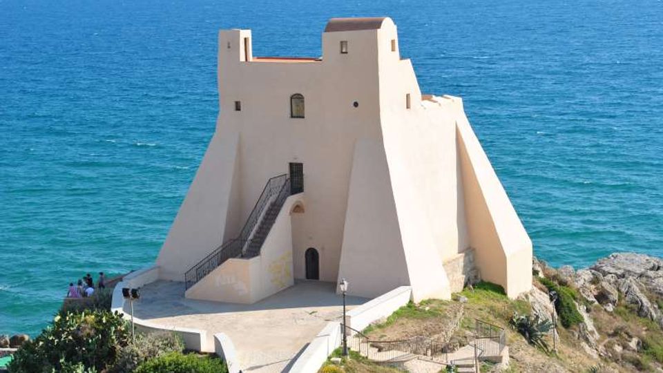 Torre costiera fortificata intonacata di bianco, con mura inclinate e scala esterna in metallo, costruita su uno sperone roccioso. Sullo sfondo si vede il mare aperto, con un piccolo piazzale e un sentiero di accesso davanti all’edificio.