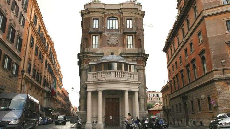 Facciata di un edificio storico in mattoni su una piazza, con grande portico ad archi e colonne e balcone semicircolare sopra l’ingresso. Ai lati si vedono due strade con palazzi alti in stile tradizionale e veicoli parcheggiati lungo i bordi.