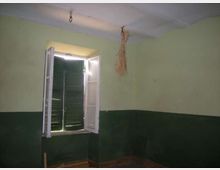 Interno di una stanza spoglia con pareti bicolori verde chiaro e verde scuro e soffitto bianco. Una finestra con persiane verdi e ante bianche aperte lascia entrare luce, con ganci e un fascio di paglia appeso al soffitto.