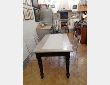 Cucina abitabile con grande tavolo rettangolare e sedie trasparenti, pavimento in cotto a spina di pesce e pareti decorate con quadri. Sul fondo si vede un camino con cappa e rivestimenti di piastrelle colorate, affiancato da elettrodomestici e piano cottura con pensili in legno.
