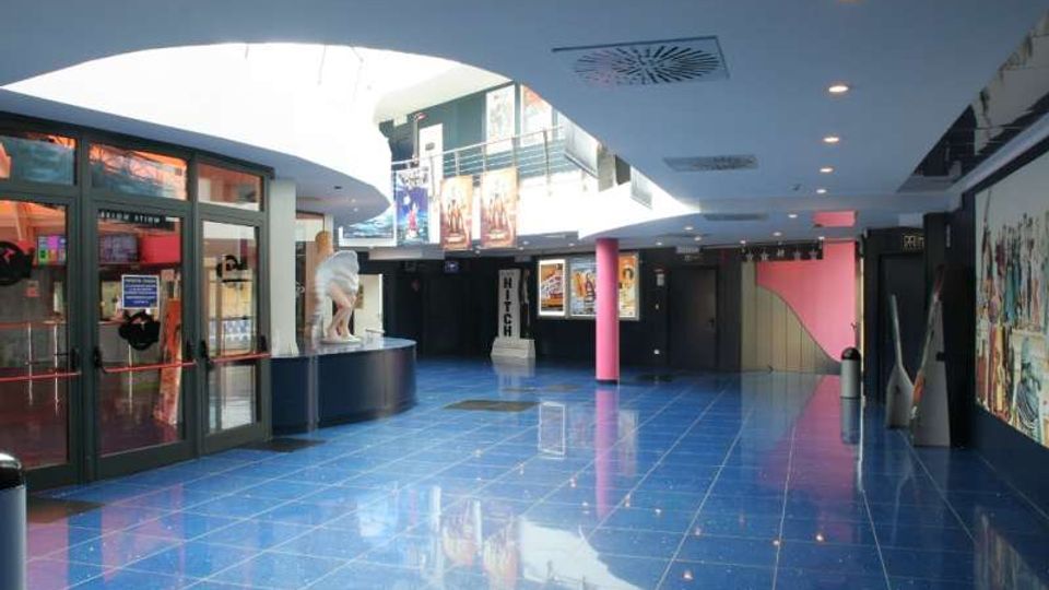 Atrio interno di un cinema o complesso commerciale con pavimento lucido in piastrelle blu, soffitto bianco curvo con faretti e lucernari. Ai lati si vedono ingressi vetrati, colonne rosa, biglietteria e pareti con poster e murales.