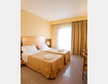 Camera d’albergo con due letti singoli affiancati, testiera in legno e biancheria chiara con asciugamani piegati. Ampia portafinestra con tende beige che lascia entrare luce naturale su pavimento piastrellato.