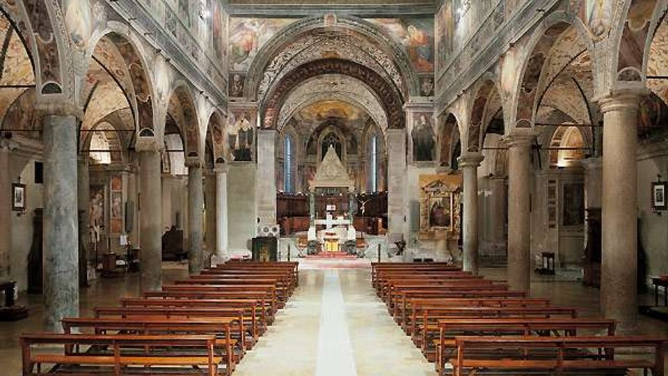 Interno di una chiesa a tre navate con file di banchi in legno lungo una navata centrale che conduce all’altare. Colonne in pietra e arcate laterali sostengono volte decorate, con affreschi sulle pareti e soffitto a cassettoni.