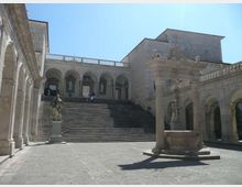 Cortile in pietra di un complesso monumentale con ampia scalinata centrale che conduce a un portico ad archi e logge superiori. In primo piano si vede una fontana in pietra e statue su piedistalli, circondate da edifici in muratura con arcate laterali.