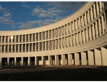 Edificio monumentale a pianta curva con lunga facciata in pietra, scandita da file regolari di alte finestre e colonne. In primo piano si vede un porticato ad archi che si affaccia su un’ampia area pavimentata, sotto un cielo parzialmente nuvoloso.