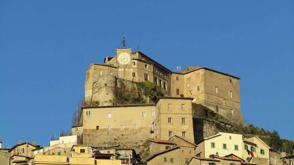 Complesso fortificato in pietra su una collina, con edifici storici sovrapposti e una torre con grande orologio sulla facciata. In basso si vede il borgo con case addossate e tetti in coppi, sullo sfondo di cielo sereno.