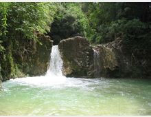 Piccola cascata che scende tra rocce e muri in pietra in una pozza d’acqua verde-azzurra. Intorno si vede una fitta vegetazione di alberi e arbusti che circonda il corso d’acqua.