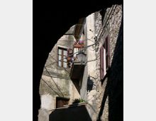 Facciate di case in pietra e intonaco in un vicolo stretto, con balconcino decorato da fiori, lampione a parete e cavi elettrici a vista. Finestre con persiane scure e muri irregolari creano un ambiente urbano storico.