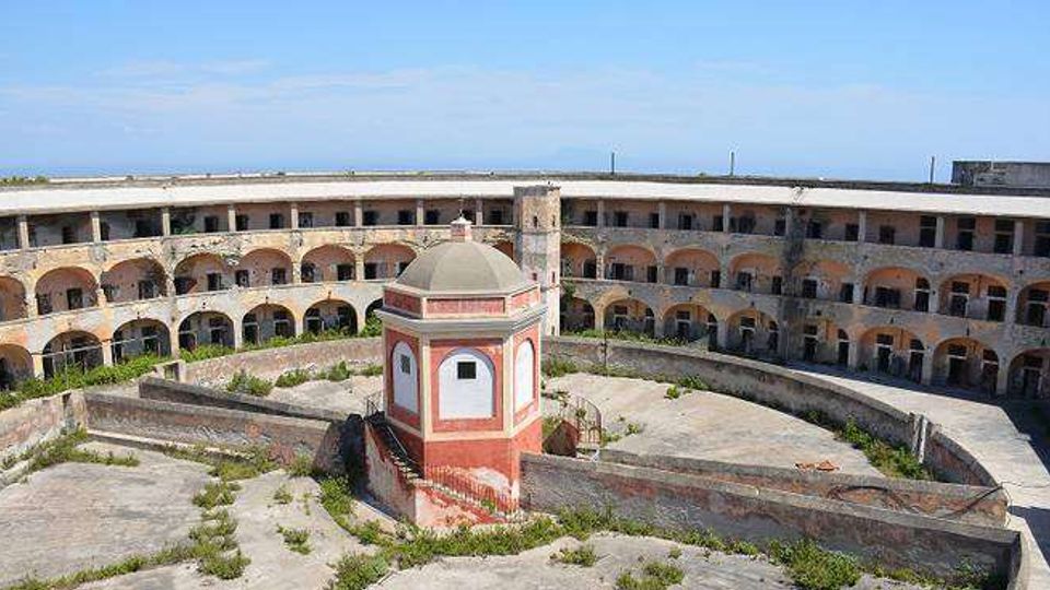 Cortile interno di una grande struttura circolare in muratura, con due livelli di porticati ad archi e finestre lungo l’anello esterno. Al centro si trova un piccolo edificio ottagonale con tetto a cupola, circondato da pavimentazione in cemento e aree con vegetazione spontanea.