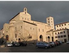 Grande chiesa in pietra in stile romanico con facciata massiccia, portali ad arco e piccole finestre, affiancata da un campanile quadrato. L’edificio si affaccia su una piazza con auto parcheggiate e palazzi adiacenti sotto un cielo scuro.