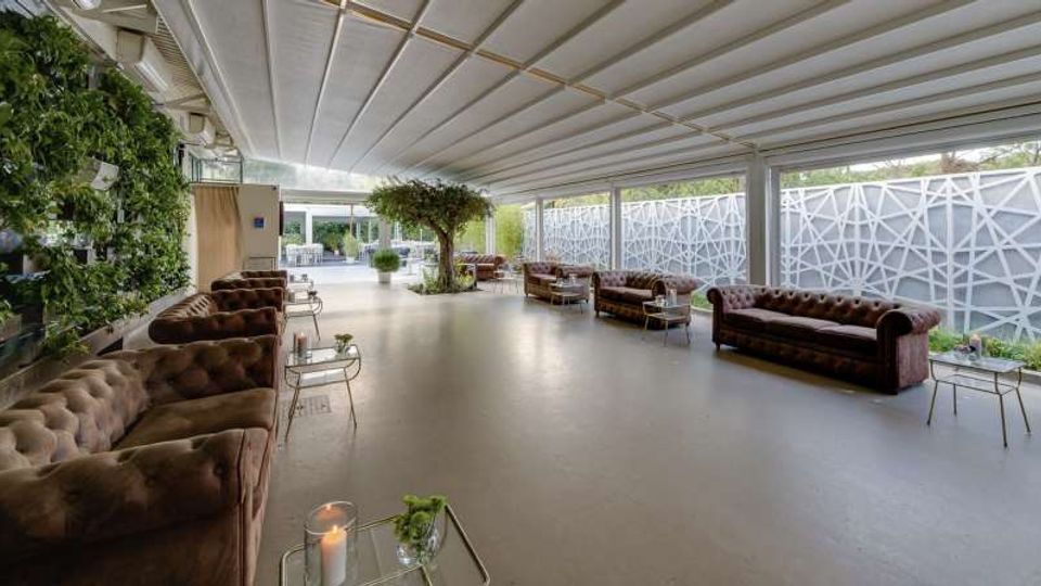 Ampia lounge moderna con soffitto a pannelli e grandi vetrate su un lato, schermate da pannelli decorativi bianchi. Divani in pelle scura con tavolini in metallo e pareti verdi con piante creano un’area relax luminosa.