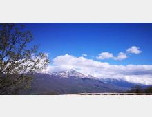Vista panoramica di una catena montuosa con cime innevate sotto un cielo azzurro con nuvole bianche. In primo piano si vedono rami di un albero sulla sinistra e una staccionata in legno lungo il bordo inferiore dell’immagine.