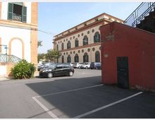 Parcheggio asfaltato con auto in sosta davanti a un edificio storico a due piani con facciata chiara, finestre ad arco e decorazioni circolari. In primo piano si vede un muro rosso con una porta e, a sinistra, parte di un edificio con persiane verdi.