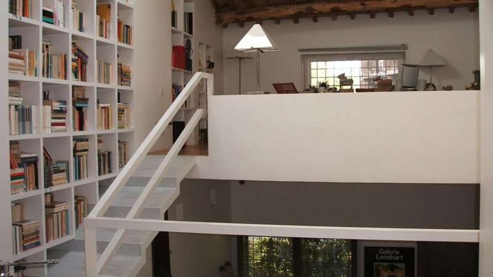 Interno di un loft su più livelli con soppalco e scala bianca, pareti chiare e grande libreria a tutta altezza piena di libri. Soffitto con travi in legno a vista, finestra sul fondo e affaccio su un livello inferiore con ampie superfici vetrate.