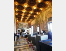 Interno di una grande sala museale con soffitto a cassettoni ottagonali illuminato e pareti affrescate con scene e architetture classiche. Nella sala sono presenti piedistalli e teche con sculture e busti, finestre alte sui lati e cordoni rossi che delimitano il percorso sul pavimento in marmo.