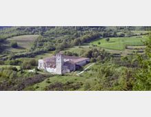 Veduta panoramica di un monastero in pietra con tetti in laterizio e torre quadrata centrale, situato in una valle. Intorno si vedono prati verdi, campi coltivati e colline boscose attraversate da una strada sterrata.