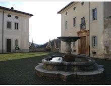 Cortile lastricato con una fontana in pietra al centro, circondato da edifici storici intonacati con finestre alte, porta monumentale e piccolo balcone in ferro battuto. Sullo sfondo si vede un muretto con elementi decorativi e un paesaggio collinare.