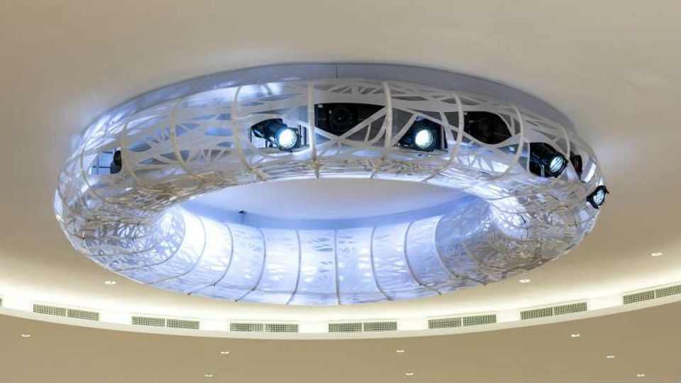 Soffitto interno moderno con grande elemento circolare sospeso, rivestito da una struttura reticolare bianca e dotato di faretti integrati. Intorno si vedono superfici curve illuminate e una fascia di piccole luci incassate nel controsoffitto.
