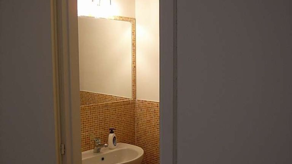 Piccolo bagno visto dalla porta, con lavabo a colonna e specchio illuminato da una lampada a parete. Le pareti sono rivestite a metà altezza con mosaico in piastrelle marroni e il resto è intonacato chiaro.