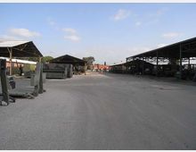 Ampio piazzale asfaltato in un’area industriale, con grandi tettoie metalliche aperte ai lati che coprono attrezzature e veicoli. Sullo sfondo si vedono altri capannoni e un cielo parzialmente nuvoloso.