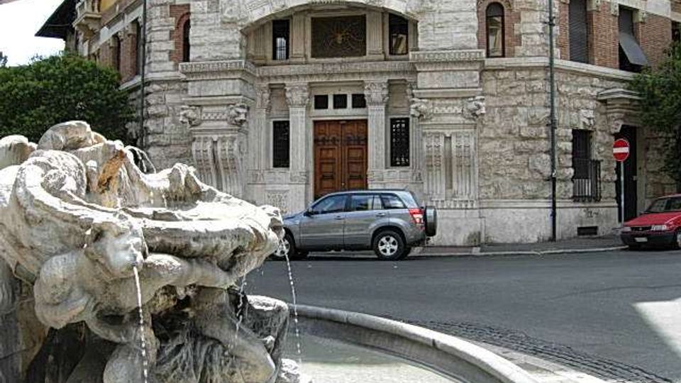 Fontana in pietra con sculture e getti d’acqua in una vasca circolare, in primo piano su una strada urbana. Sullo sfondo si vede la facciata di un edificio storico con portale in legno, decorazioni in pietra e finestre ad arco.