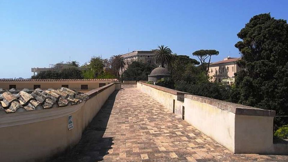 Lungo camminamento sopraelevato in pietra, affiancato da muretti chiari con copertura in coppi su un lato. Sullo sfondo si vedono edifici bassi e alberi mediterranei, tra cui palme, sotto un cielo sereno.