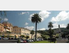 Ampia piazza urbana con parcheggio e prati curati, fiancheggiata da alti palmeti e da edifici storici color pastello con archi al piano terra. Sullo sfondo si vede una collina verde e, a destra, una fontana monumentale con statua sotto un cielo parzialmente nuvoloso.