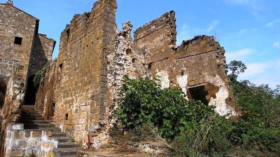Rovine di un edificio in pietra con muri parzialmente crollati e intonaco consumato, affiancate da una scalinata in pietra. Vegetazione fitta cresce alla base e all’interno delle strutture, con cielo azzurro sullo sfondo.